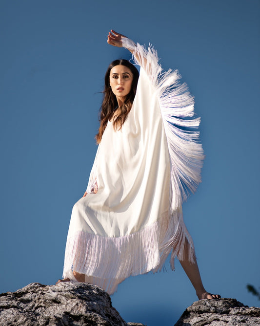Woman in a white dress with fringe standing on rocks against a clear blue sky