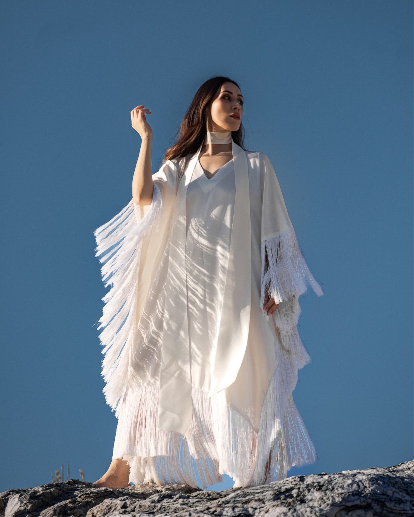 Woman in a white dress with fringe standing on a rocky surface against a clear blue sky.